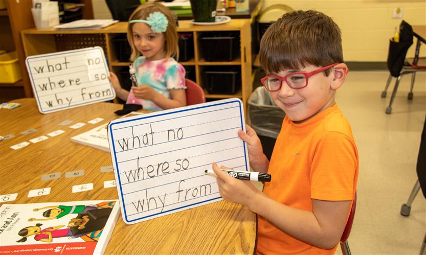 A boy with glasses and wearing an orange shirt writes on a dry erase board.